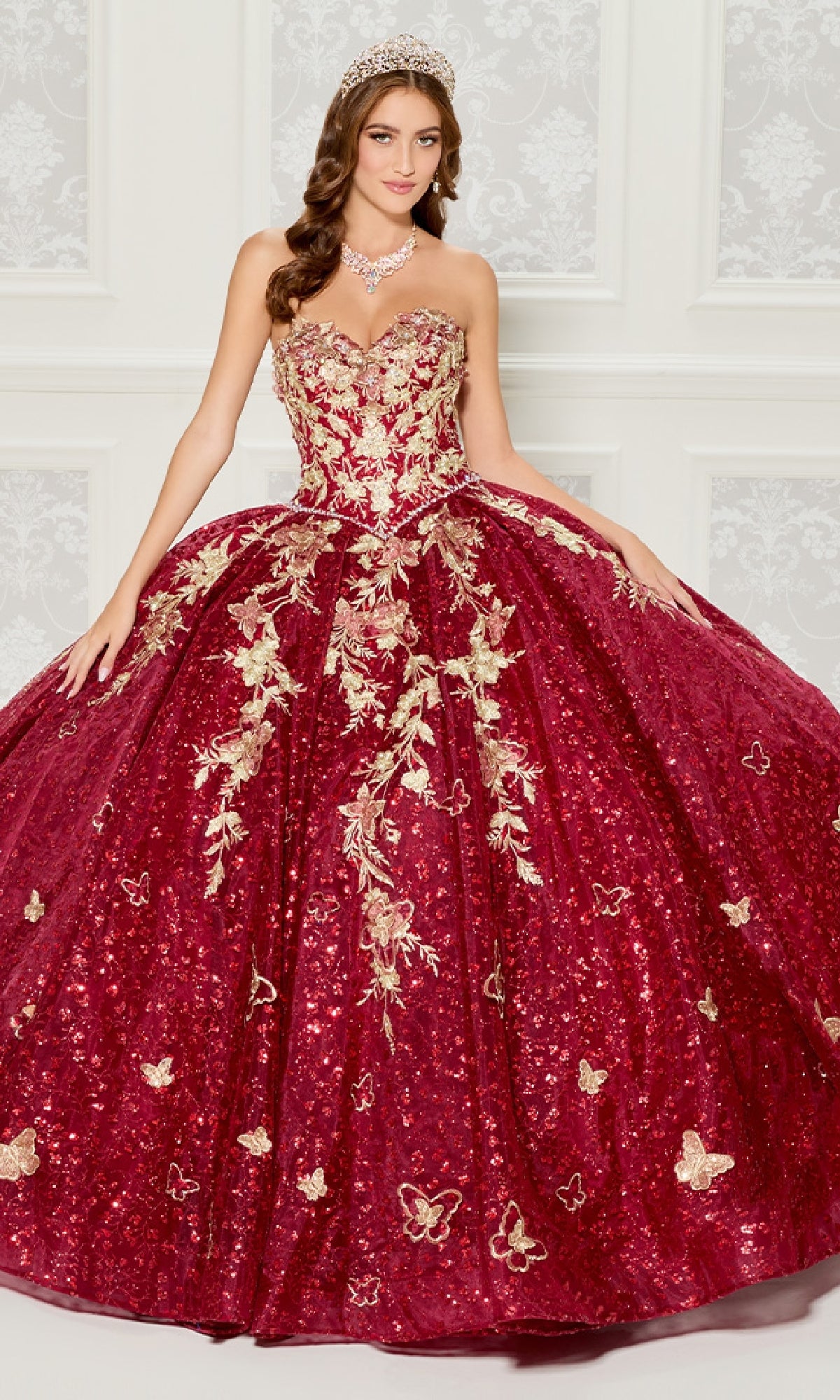 A woman in the Strapless Quinceanera Dress: Princesa PR30121, with red and gold embroidery and a full skirt, stands before a white patterned wall wearing a tiara and necklace.